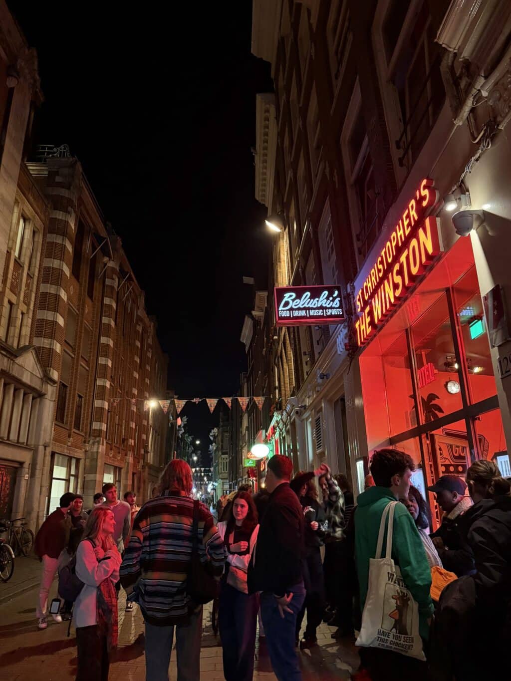 Night view of Flying Pig Uptown hostel on a lively street with groups of people and colourful signag.