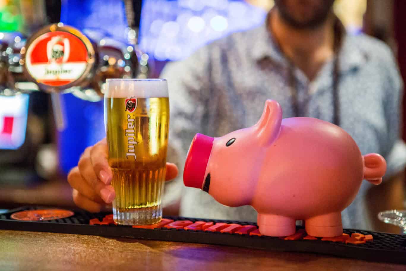 Person holding a beer glass next to a pink piggy bank at Flying Pig Uptown Hostel bar.