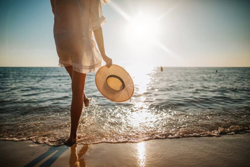 Beach sunset with woman in white dress holding straw hat.