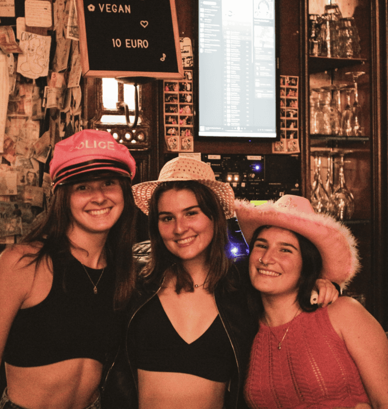 Girls in colourful hats smiling at the bar in a lively hostel setting.