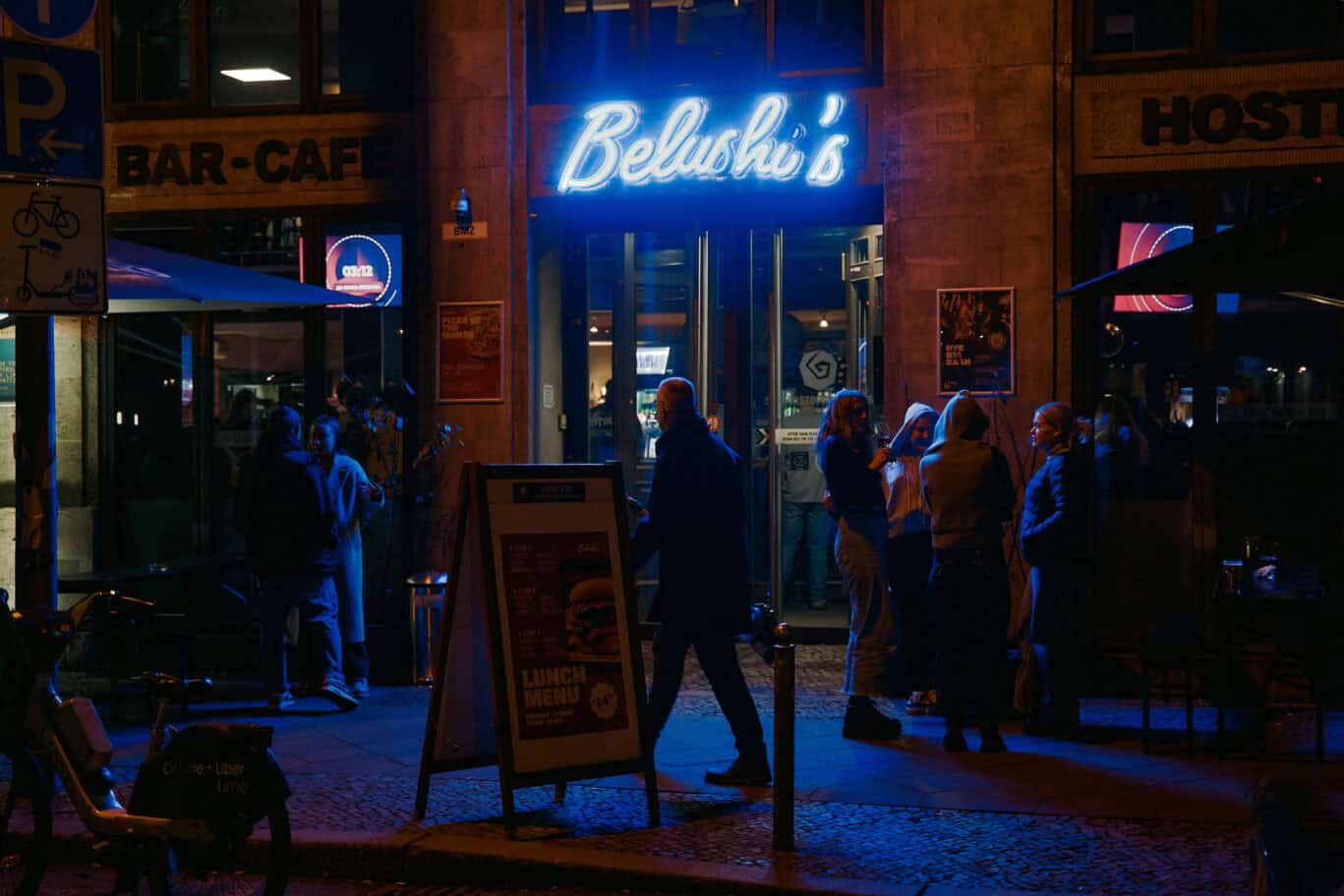 Neon sign of Belushi's bar outside at night, with people gathered nearby.