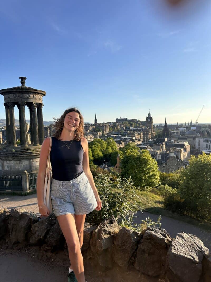 A woman standing near a historic monument with a city skyline in the background.
