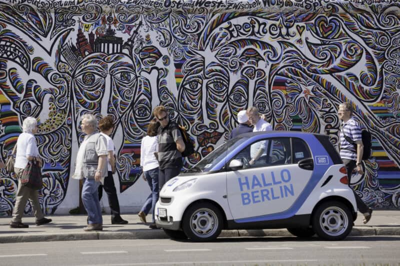 Tourists walking past a colourful mural in Berlin with a small electric car in the foreground.