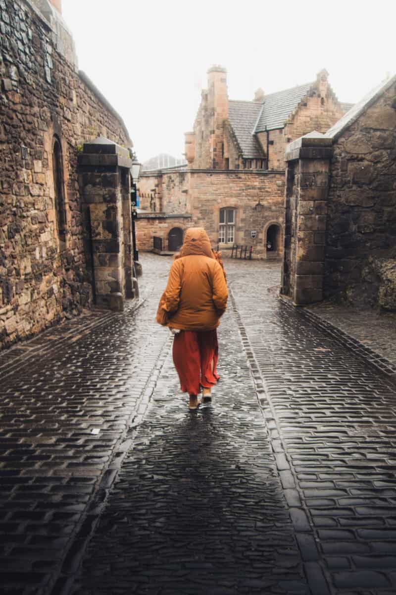 A person walking through a castle entrance on a rainy day, cobbled street and historic stone walls.