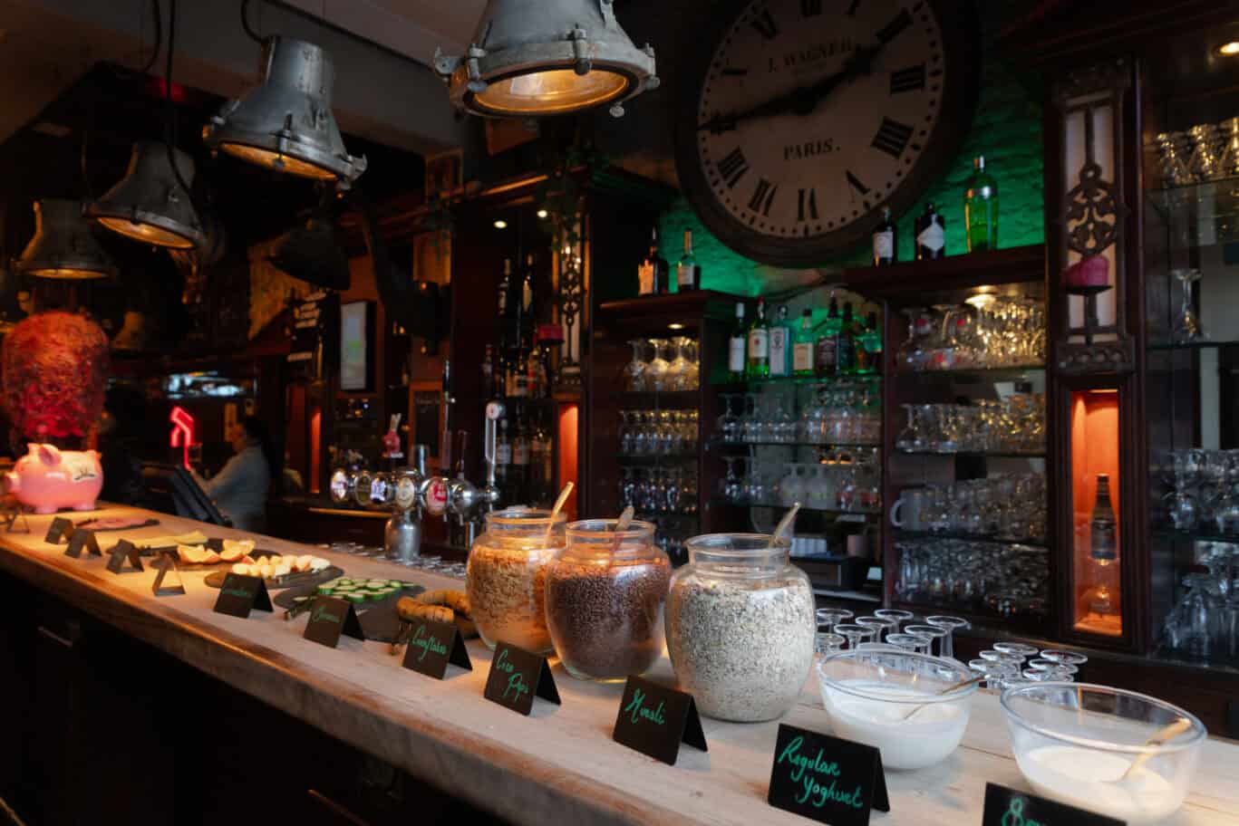 Interior view of the bar at St Christopher’s Bruges, showcasing a variety of drinks and a large cloc.
