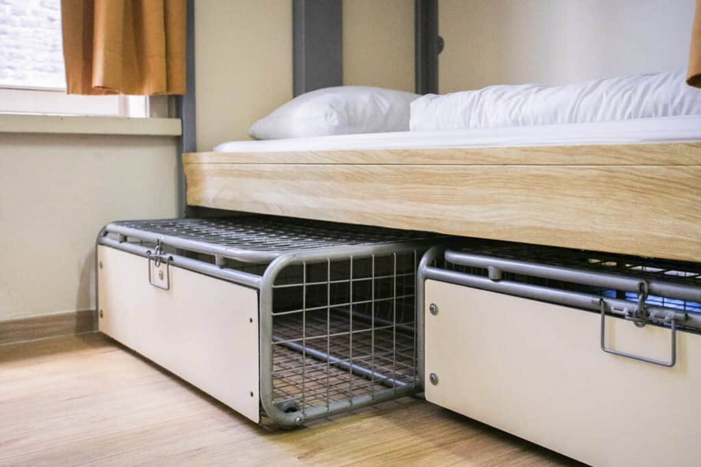 Hostel bed with storage and metal cage in a tidy, modern dorm room.