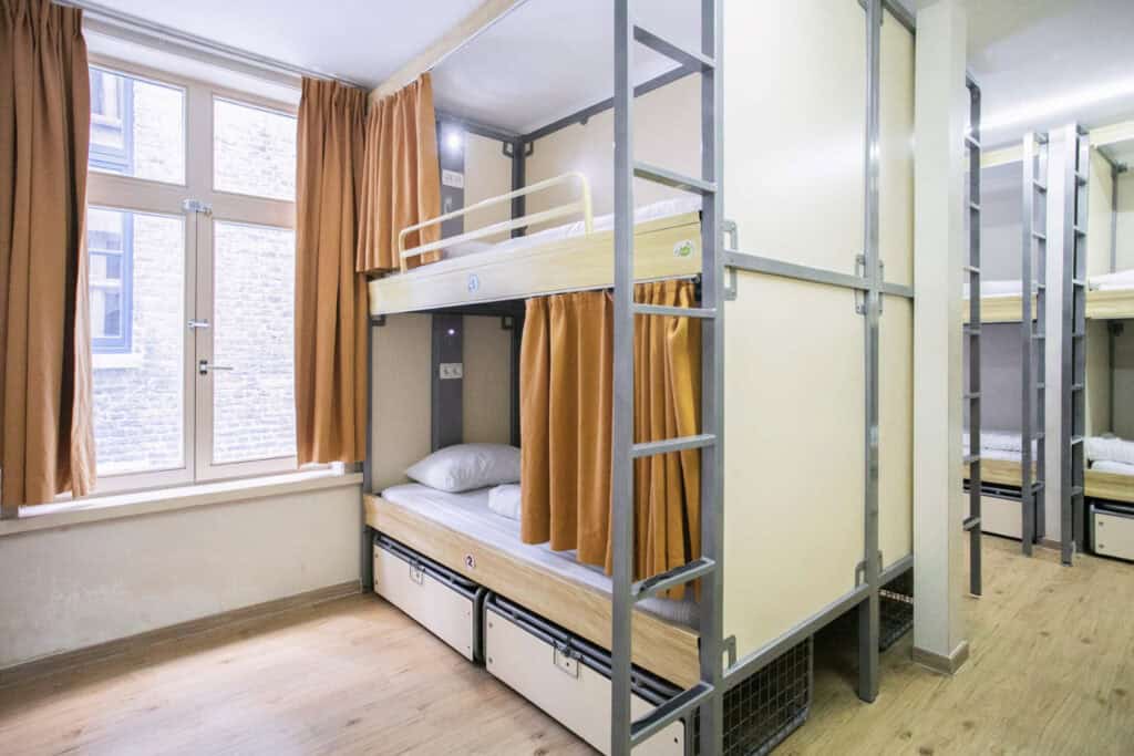 Bunk beds in a bright hostel room with large windows and orange curtains at St Christopher’s Bruges.