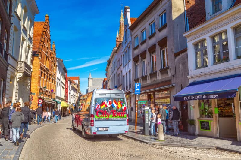 Tour bus on a lively Bruges street with shops and tourists.