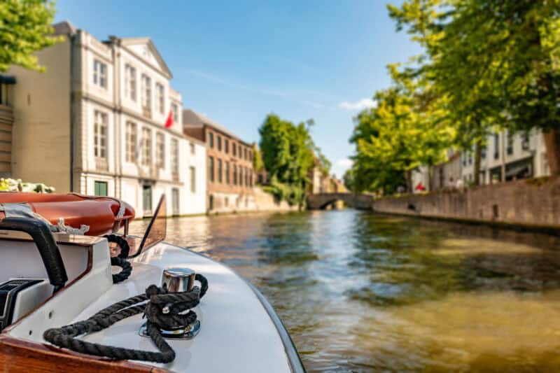 A boat on the Bruges canal with historic buildings and lush trees in the background.