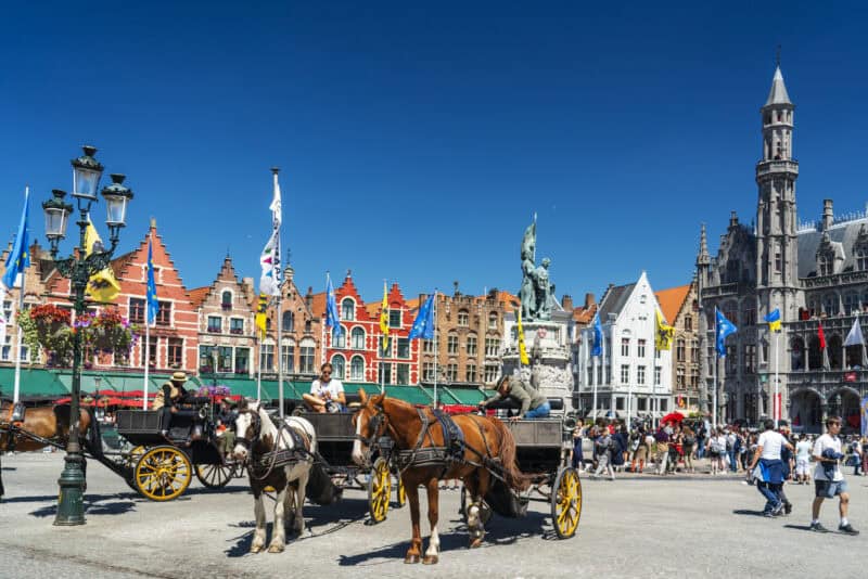 Historic Bruges city square with horse-drawn carriages and colourful medieval buildings.