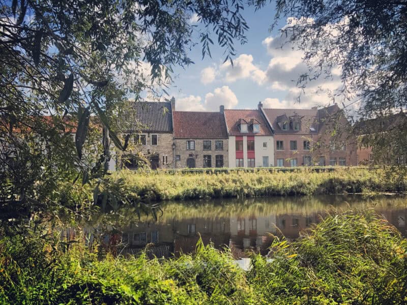 Scenic view of Bruges canal with historic buildings and lush greenery under a blue sky.