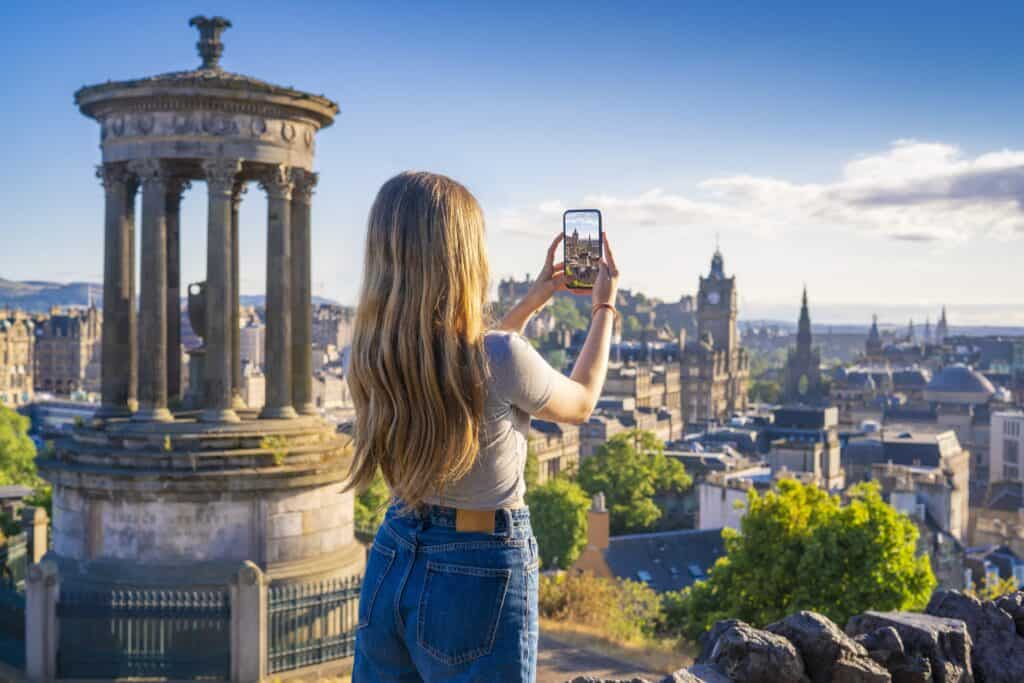 Woman taking a photo of Edinburgh skyline from Calton Hill with historic monuments.