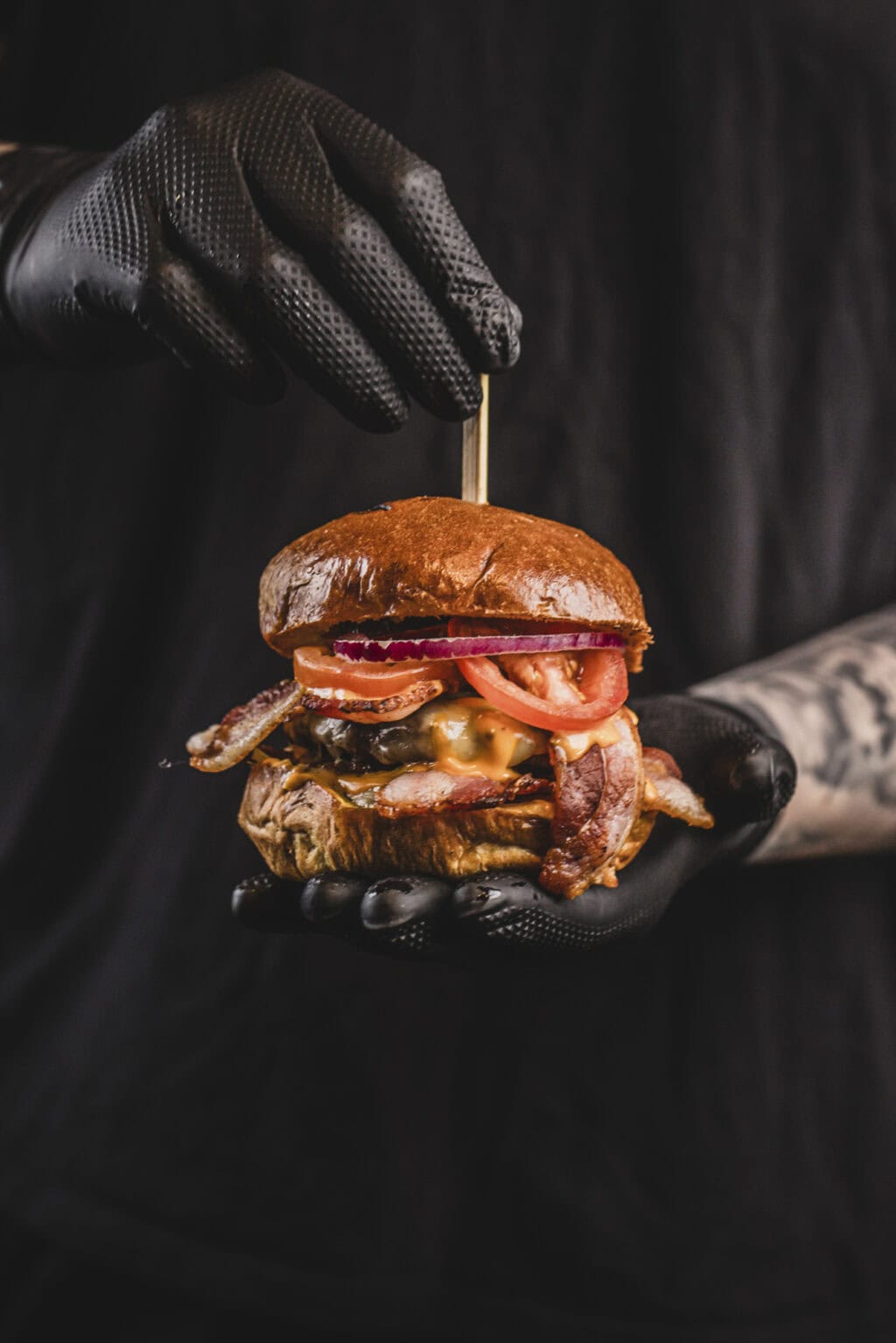Delicious bacon cheeseburger with fresh toppings held by a person wearing black gloves.