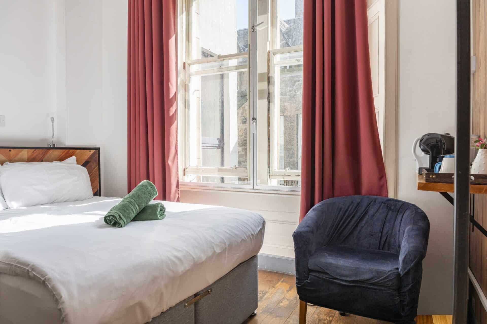 Comfortable hostel room with a large bed, green towels, and a navy armchair near a window with red c.
