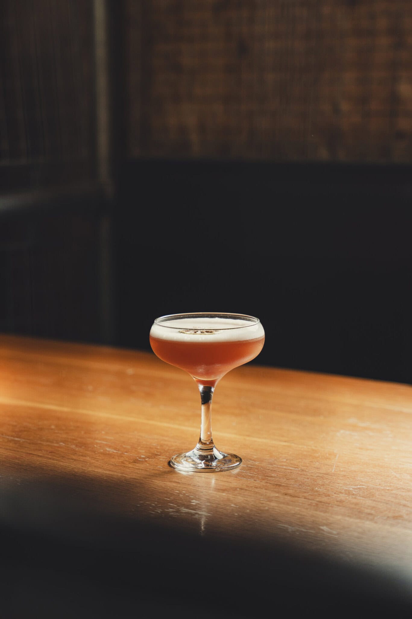 A cocktail in a coupe glass placed on a wooden surface, with a dark, cozy background.