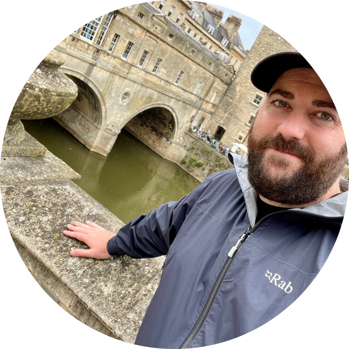 A bearded man stands beside a bridge over a river