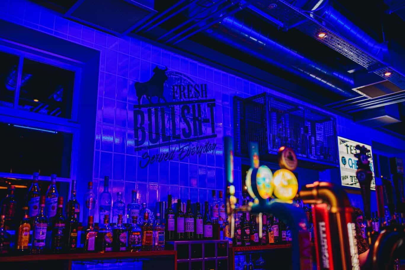 Bar area at St Christopher’s Edinburgh hostel with colourful lighting and a variety of bottles on di.