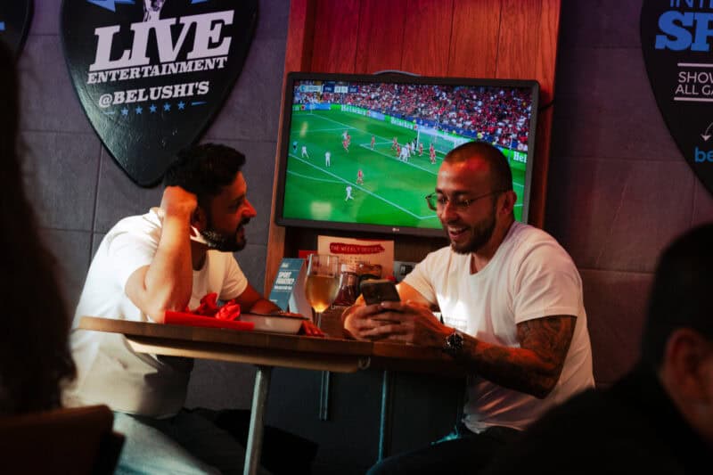 Two men enjoying drinks and watching a football match at St Christopher’s Edinburgh hostel.