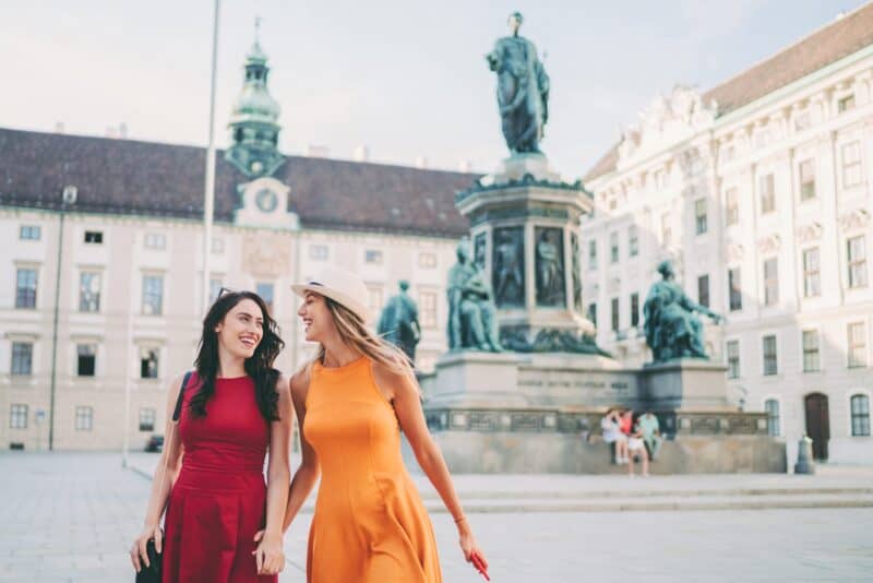 Two women in colourful dresses walking in a city square with historic buildings and statues.