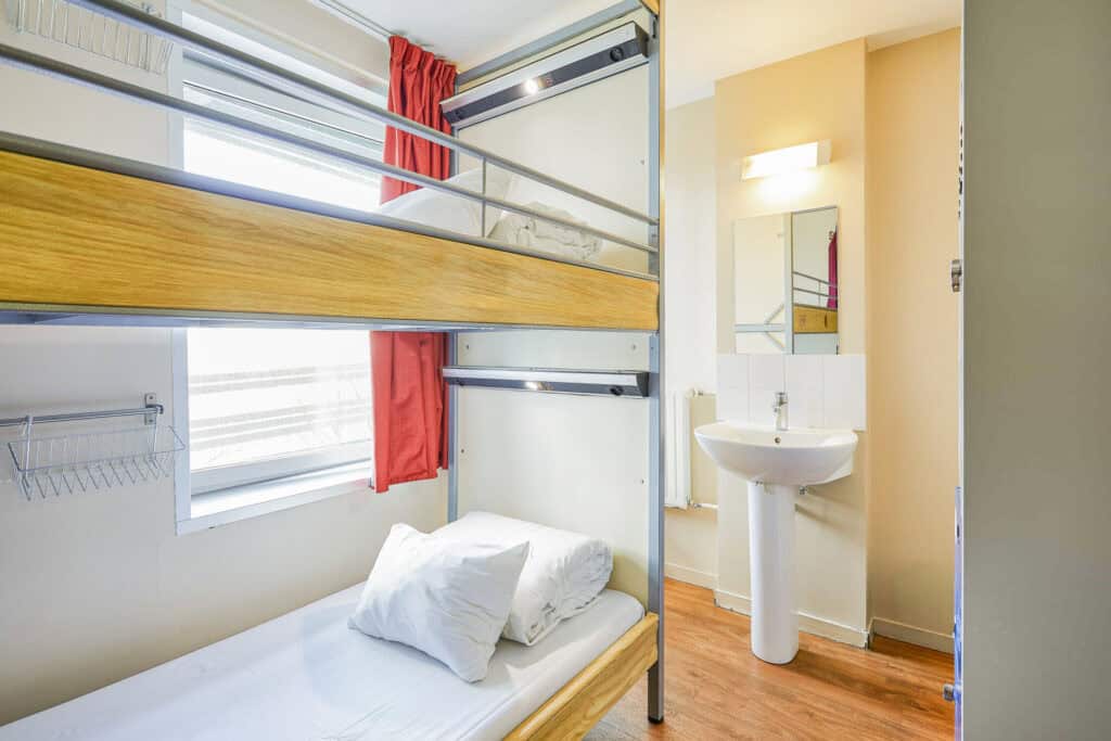 Bunk bed and sink in compact hostel room, bright with natural light and colourful curtains.