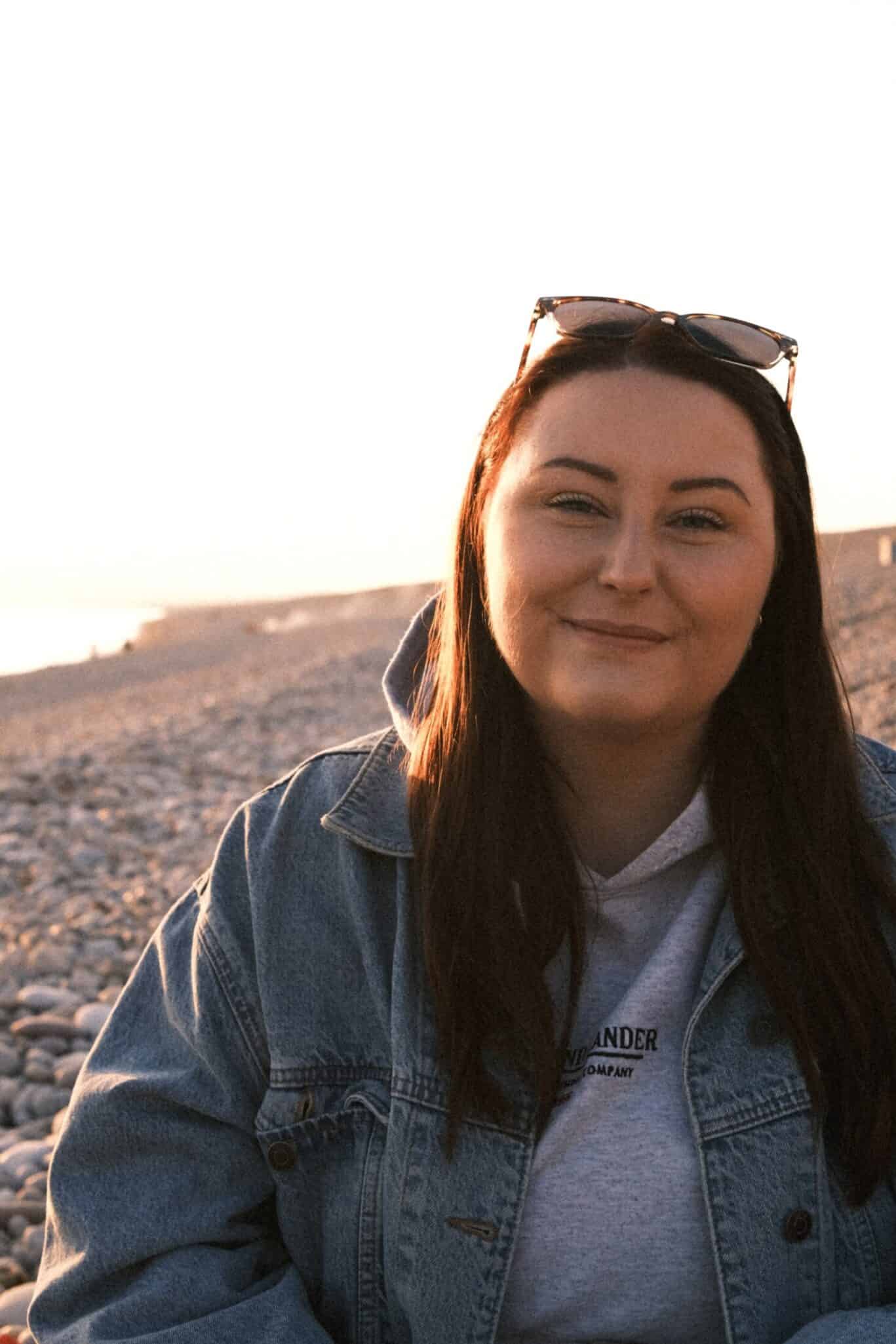 Woman smiling at the beach during sunset, wearing sunglasses and denim jacket.