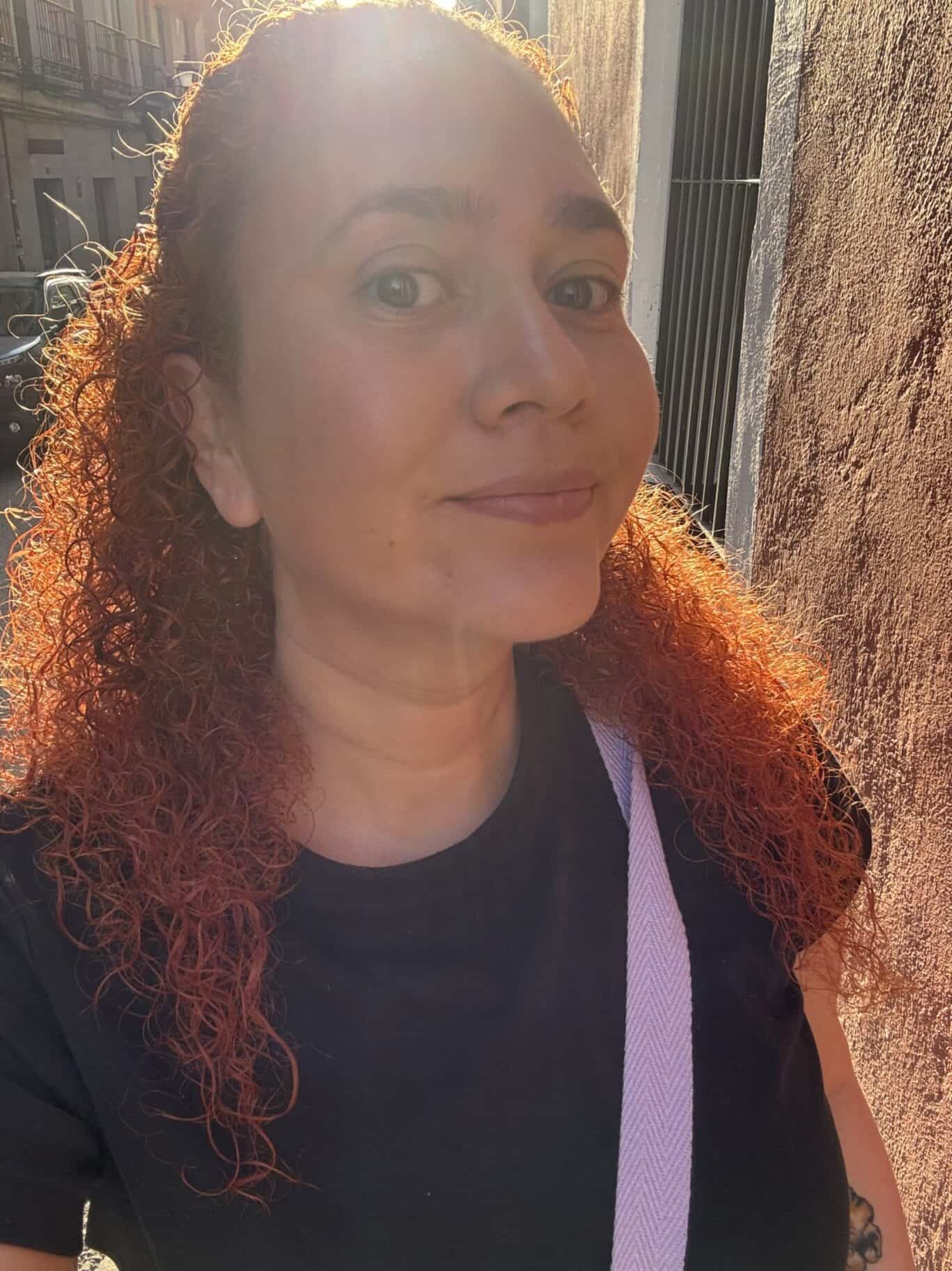 Woman with curly red hair smiling outdoors in sunlight, urban background.