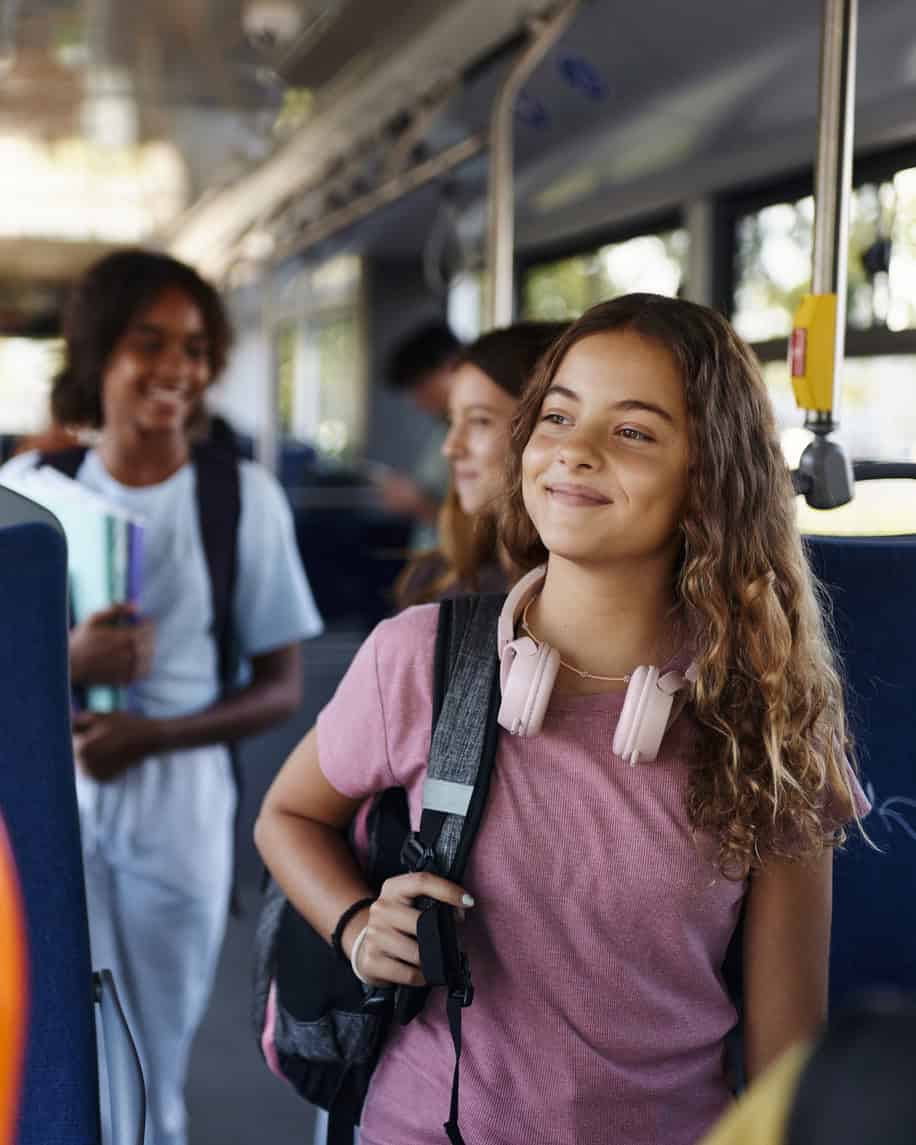 Happy schoolgirl with headphones and backpack on bus, smiling.