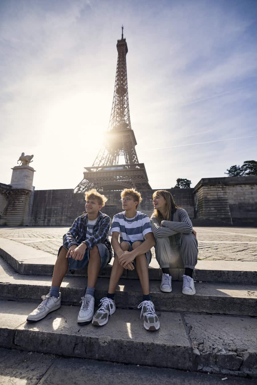 Three young people sitting on steps near Eiffel Tower in Paris, enjoying the sunny day.