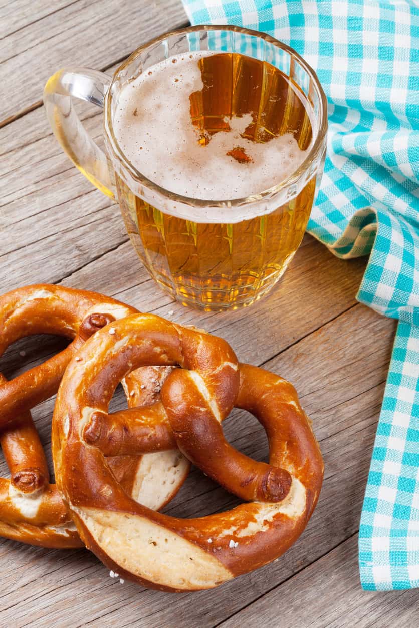 Glass of beer with pretzels on a wooden table, blue checkered cloth nearby.